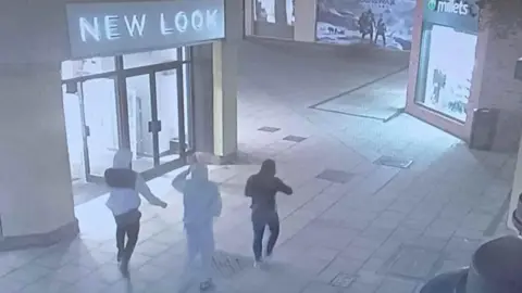 A still from CCTV showing three people in hoodies walking away from the camera with shops either side of them. One is in a white and black tracksuit, another is in all grey and another is in all black.