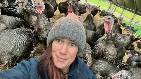 Kate Postance of Postance Poultry takes a selfie in the the purpose-built tunnel housing her turkeys. The turkeys can be seen behind her. Kate has long brown hair and is wearing a grey, woollen hat and a blue fleece.