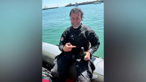 Stefan Panis A man with dark hair, swept back, holding a black bottle. He is wearing a wet-suit and sitting on the side of a boat. He has a grey beard and is smiling at the camera, pointing at the bottle.