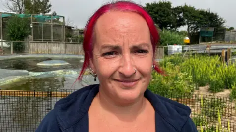 BBC News A woman wearing a blue jacket looks at the camera. She has a gold pendant necklace and red hair. She is standing in front of a pond with bushes lining one side. 