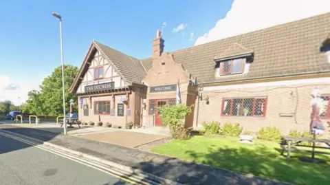 A pub with black signage outside and a lamppost. The pub has a small front garden.