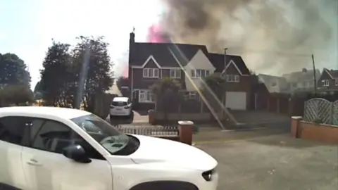 Smoke and flames are rising above a house with a white car parked on the drive.