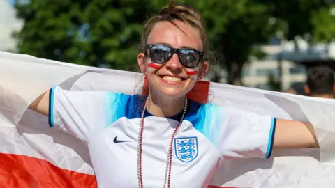 A woman in an England shirt and sunglasses smiles.