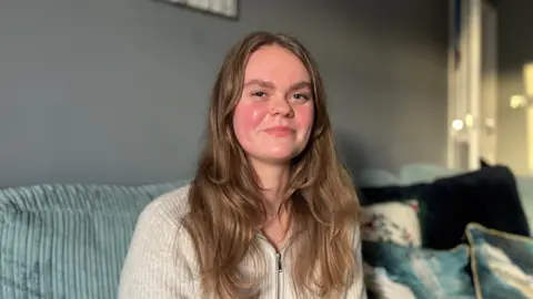 Mollie Penfold who has brown long hair and is wearing a grey quarter zip wooly jumper sitting on the sofa of her living room