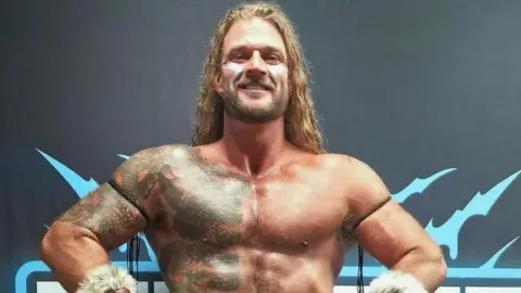 Johnny Jupiter Johnny Jupiter has long blonde hair, a beard, a large tattoo on the right of his bare torso. He is standing up and smiling, with his hands on his hips. He is in front of a black wall.