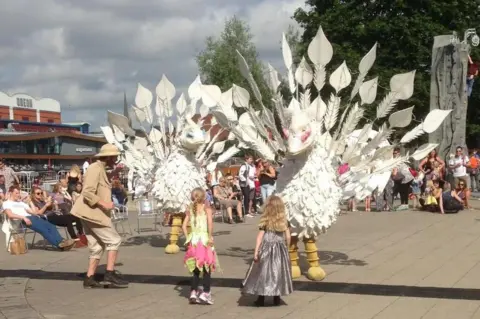 Apus Productions Tow large creatures resembling gigantic birds, with white plumage and elaborate tail feathers, stand in a busy plaza as two young girls, with their backs to the camera, watch them.
