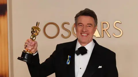 Peter Straughan poses with the Best Adapted Screenplay for Conclave award in the Oscars photo room. He is wearing a black suit and has blue and yellow ribbons pinned to his lapel.