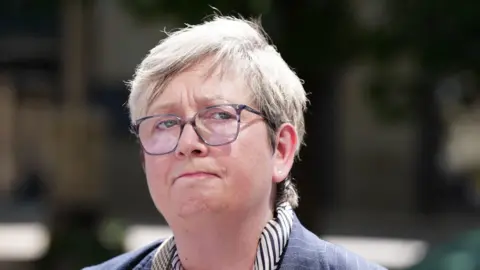 PA Media Close-up outdoor portrait of a Joanna Cherry who has short light-coloured hair wearing large, round, translucent-framed glasses, a small stud earring, and a collared shirt with narrow stripes under a textured jacket. The background is softly blurred, suggesting greenery and buildings, with bright natural sunlight illuminating the face.