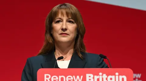 BBC Chancellor of the Exchequer Rachel Reeves, wearing a dark suit and white blouse, is standing in front of a red background. She is behind a lecturn that bears the slogan: Renew Britain.