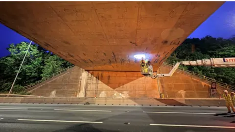 National Highways Inspectors wearing high-vis and on the end of a cherry picker, assess some of the defects under the concrete bridge over a closed M42 motorway