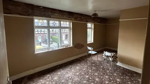 Room inside house with brick damage, patterned carpets and two chaits in the centre of the room. 