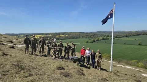 Helen Roberts A group of people, some in army gear, smile widely on a hillside next to a white pole with an Australian flag.