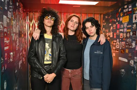 Muna stand inside a lift covered in stickers, each showing the logo of a different band. Singer Katie Gavin is in the middle, with her arms draped around her bandmates.