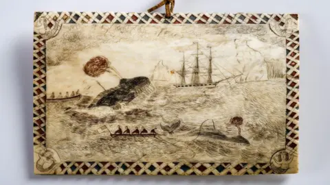A carved whale bone featuring a scene of sailors in boats hunting a whale which is leaping from the sea with a sailing ship in the background amid icebergs