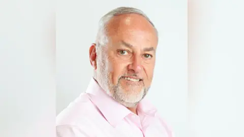 Portrait photo of councillor Mark Howard. He is wearing a pink shirt and has combed, white hair and a white beard. He is smiling.