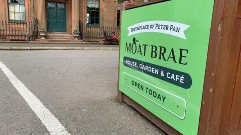 A sign for the Moat Brae visitor centre describing it as the birthplace of Peter Pan sits outside the doorway of a sandstone building