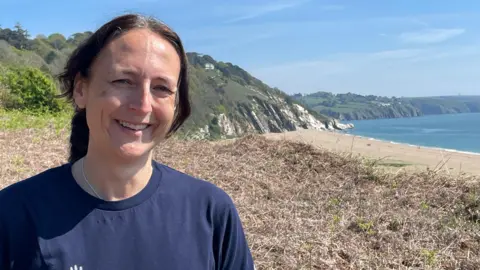 A woman in a blue t-shirt smiles at the camera with the Devon coastline and sea behind her.