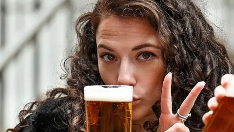 A woman and a man sitting outdoors holding pints of Lager. She is making a peace sign and he is drinking, in a casual seating area with steps and railings in the background