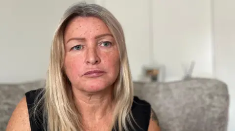 A woman in her 40s with blonde shoulder-lenght hair. She had freckles and blue eyes and a neutral, slightly sad expression. She is wearing a black top and has a tattoo on her arm. The background is blurred but appears to be a living room.