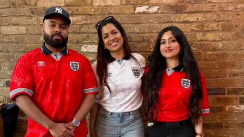 Shabna Zaheer Three people posing together in front of a brick wall, with the man and woman on the right wearing red and white England football shirts.