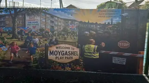 Banner on a play park fence, showing children playing sunshine and men being rejected at a barrier. 