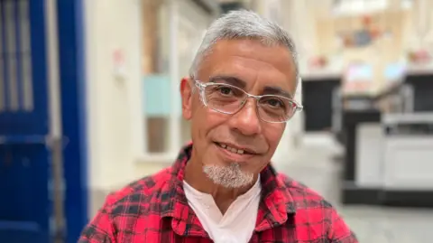 A brown skinned man with grey hair and a goatee beard looking towards the camera. He's wearing glasses and a red and black checked shirt. 