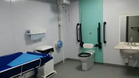 An accessible changing place facility, with a hoist, a bed, toilet and sink. It is a white room with black hand rails to improve accessibility.
