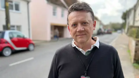 Stuart Woodward/BBC Councillor Paul Thorogood stands on a pavement with houses and parked vehicles behind him. He is wearing a dark v-neck jumper over a white open-necked shirt, and has a black lanyard around his neck. Paul has short dark hair and is looking at the camera.