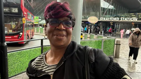 A woman with short red hair wearing a striped top and hoodie stands in front of Stratford Station entrance.