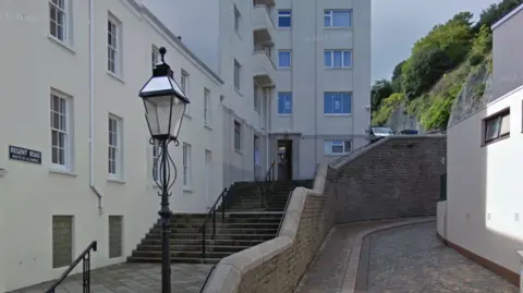A set of stone steps leading up to a door for the Regent House. There is a sign on the wall that reads REGENT ROAD and underneath RUETTE DE LA COMEDIE. To the right of the steps is a wall and a path next to a building. There is an ornate iron work lamp post in the foreground.