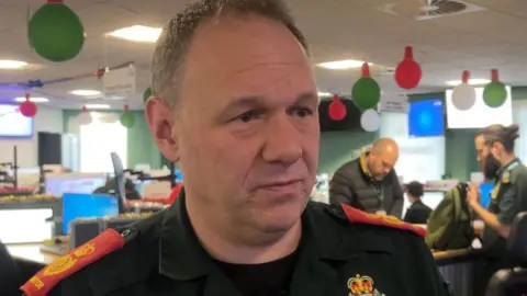 Dr Matt Thomas in a control room. He is being interviewed and is looking away from the camera. People and computers are visible in the background, with Christmas decorations hanging from the ceiling. 