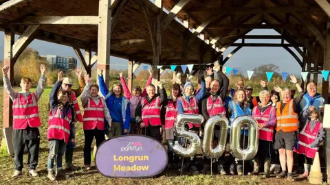 Parkrun A row of 19 people, mostly in pink hi-vis jackets have their hands in the air and are smiling at the camera. They're stood underneath a barn-like structure, and in front of them is a blue sign which says 'Longrun Meadow'.