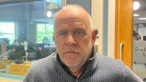 An older man with blue eyes and grey hair and stubble looks into the camera. He is wearing a grey quarter zip. In the background is a studio.