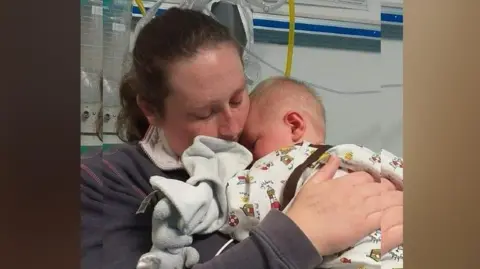 family handout Beth holding Riley as a baby in hospital. She is wearing a blue jacket and he is snuggled asleep against her. 