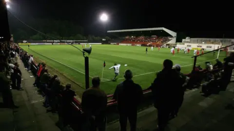 Getty Images Kettering Town's Rockingham Road stadium during a evening match. A crowd is watching on as a player in a white shirt takes a corner kick.