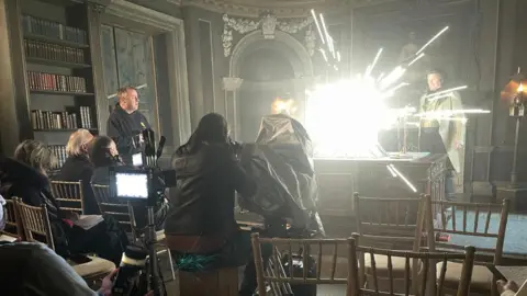 Frank Corr An interior film set inside an ornate, historic room with decorative arches, carved wall details, and shelves of books. In the foreground, rows of wooden chairs face a camera setup surrounded by crew members and filming equipment. At the centre of the scene, a bright burst of light and sparks erupts from a device on a desk as part of a staged effect, illuminating the room. Harrison in period‑style clothing stands nearby while the crew observe and film.