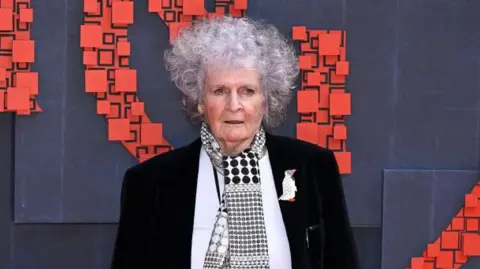Getty Images Maggi Hambling attends the National Portrait Gallery Re-Opening at National Portrait Gallery on June 20, 2023 in London. She has short, curly grey hair and wears a black jacket with a white top underneath and a black and white scarf with circles on it. 