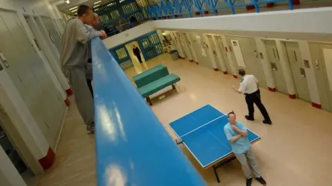 Prisoners leaning over a balcony inside the prison. Below them, an inmate is leaning on a blue table tennis table as a guard walks past.