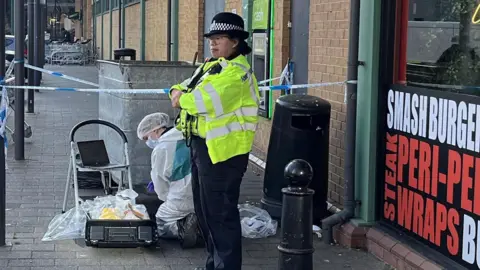 A police officer wearing a yellow stands with folded arms outside a fast food shop. Kneeling to her right is a crime scene investigator dress in a white suit.