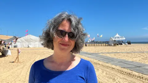 GUY CAMPBELL/BBC Festival organiser wearing a blue top and sunglasses standing on sandy beach