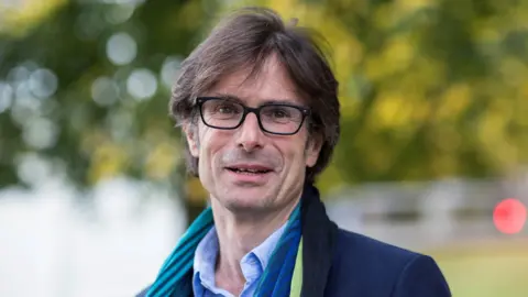 ITV political editor Robert Peston looks at the camera, with some greenery blurred behind him. He has long brown hair and is wearing glasses. A scarf can just be seen around his neck, and he is wearing a shit blue shirt, and navy coat. 