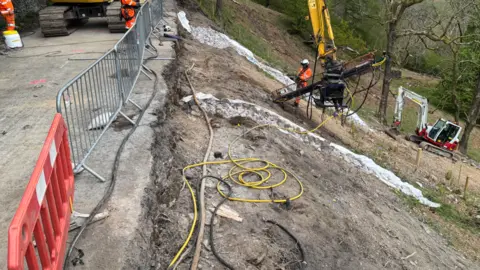 A large digger installs a metal rod in the slope near Kirkstone Pass. The yellow digger is on the road and three people wearing orange fluorescent uniform watch over. There are silver gates separating the road from the slope and a smaller digger lower down on the slope.