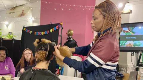 A woman with braided afro hair styles a woman with afro hair in a room, watched by an older woman with short afro hair and a young woman with long red hair.