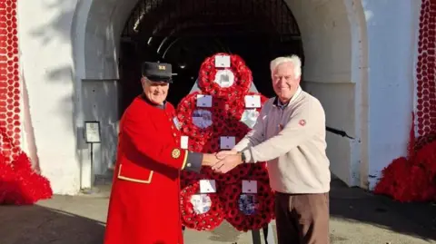 Two men including Chelsea Pensioner Michael Gue are stood in front of the tunnel which is covered in poppies and two large Jersey flags They are shaking hands. The poppies are small and all handmade. They are all stood next to a large triangular wreath made up of poppies.