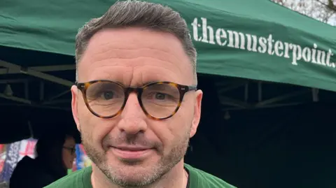 Nicola Haseler/BBC A man wearing glasses and a green t shirt smiles slightly as he looks into the camera, standing in front of a green gazebo bearing the words the muster point