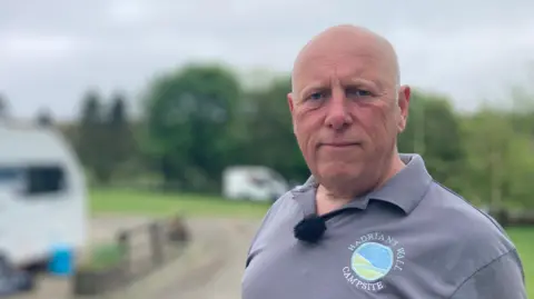 BBC Steve Miller is standing on his campsite and looking at the camera. He is bald and is wearing a grey polo shirt with a "Hadrians Wall Campsite" logo on it.