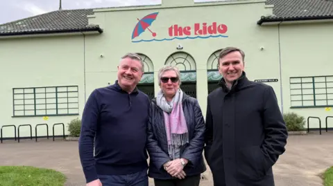 Andrew Pakes A man wearing a navy knitted top standing next to a woman wearing a blue padded jacket and a pink and grey scarf. On the right is a man wearing a black coat with his arms in his pockets. Behind them is the outside wall of the lido, with the words "The Lido" written on it in red