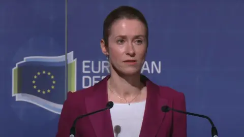 EEAS Kaja Kallas pictured in front of a podium wearing a white shirt and a purple suit jacket. There are two microphones pointing at her.