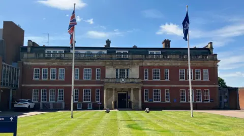 A large brick building with bay windows. A lawn is in front of the building with two flags - the Union Jack and Northamptonshire Police logo - flying.