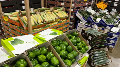 Shaun Peel/BBC Boxes of fresh produce inside Fountain Fresh's warehouse. In the foreground are four boxes of green peppers, propped up, as if they were on a market stall. Next to them are boxes of cucumbers, with courgettes behind them. Further back on the left are corns on the cob, with a box of yellow peppers in the top right corner.